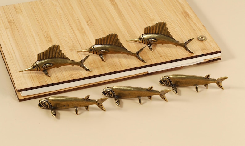 Op de afbeelding: Bronskleurige decoratieve hardware in de vorm van vissen. De afbeelding toont zes vissen, drie met hoge rugvinnen en drie met kortere vinnen, gerangschikt op een houten oppervlak.