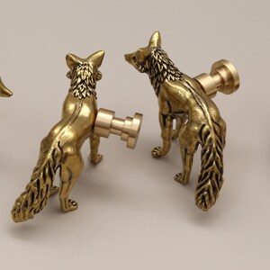 Op de afbeelding: Drie messingkleurige kastknoppen in de vorm van een vos. De knoppen zijn gedetailleerd met getextureerde vacht en een staande pose. De vossenknoppen hebben een warme goudkleur en zijn gemonteerd op een lichtgekleurd oppervlak. Een houten schijf staat op de achtergrond.