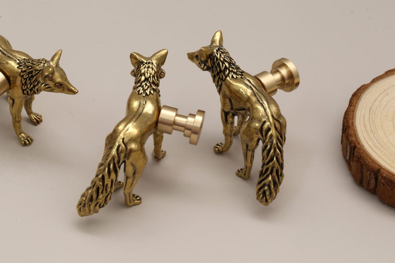 Op de afbeelding: Drie messingkleurige kastknoppen in de vorm van een vos. De knoppen zijn gedetailleerd met getextureerde vacht en een staande pose. De vossenknoppen hebben een warme goudkleur en zijn gemonteerd op een lichtgekleurd oppervlak. Een houten schijf staat op de achtergrond.