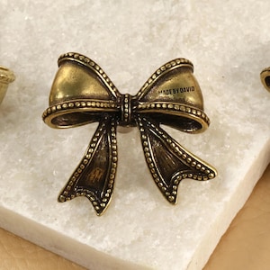 Puede incluir: Primer plano de tres herrajes decorativos en forma de lazo de color oro antiguo. El lazo central tiene el texto "MADE BY DAVID". Los lazos están sobre una superficie de mármol blanco.