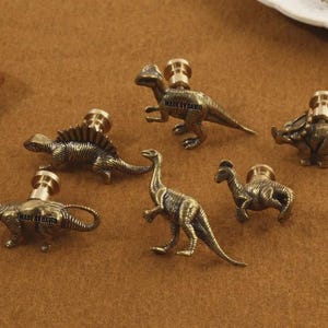 May include: Six antique brass dinosaur cabinet knobs in various poses. Each knob features a different dinosaur design, including a Stegosaurus, a Brontosaurus, and a Triceratops. The knobs are mounted on a brown surface.