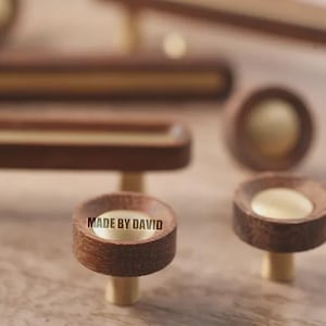 Puede incluir: Colección de herrajes de madera para armarios, que incluye pomos y tiradores, con detalles en latón. Los pomos tienen el texto "MADE BY DAVID". Los herrajes son de color marrón cálido, con un diseño moderno.