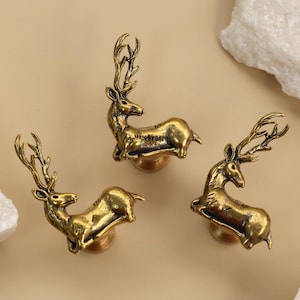 Puede incluir: Tres tiradores de armario de latón con forma de ciervo. Cada tirador presenta un diseño detallado de ciervo con cornamenta prominente. Los tiradores son de color dorado y están colocados sobre un fondo neutro con detalles de piedra blanca.