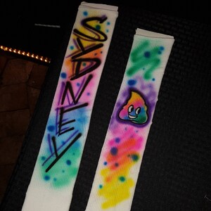 May include: Pair of white socks with airbrushed designs. One sock features the name "SANEY" in black and colorful spray paint. The other sock has a smiling poop emoji with colorful paint splatters.