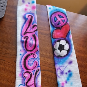 May include: Two white athletic socks with airbrushed designs. One sock features the name "LUCY" in pink and black, outlined in blue. The other sock has a peace sign, a red heart, and a soccer ball, with blue and pink accents.