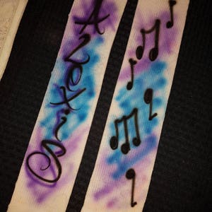 May include: Two white socks with airbrushed designs. One sock features the word "Axton" in black script over a blue and purple background. The other sock has black musical notes over a similar background.