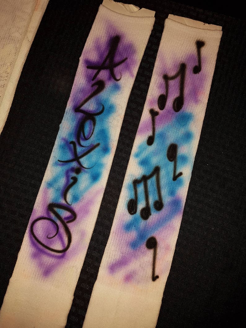 May include: Two white socks with airbrushed designs. One sock features the word "Axton" in black script over a blue and purple background. The other sock has black musical notes over a similar background.