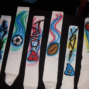 May include: White athletic socks with airbrushed designs. Each sock features a name, sports ball, or cartoon character. Designs include a soccer ball, football, basketball, and a smiling blue character. The names are in black graffiti-style lettering.