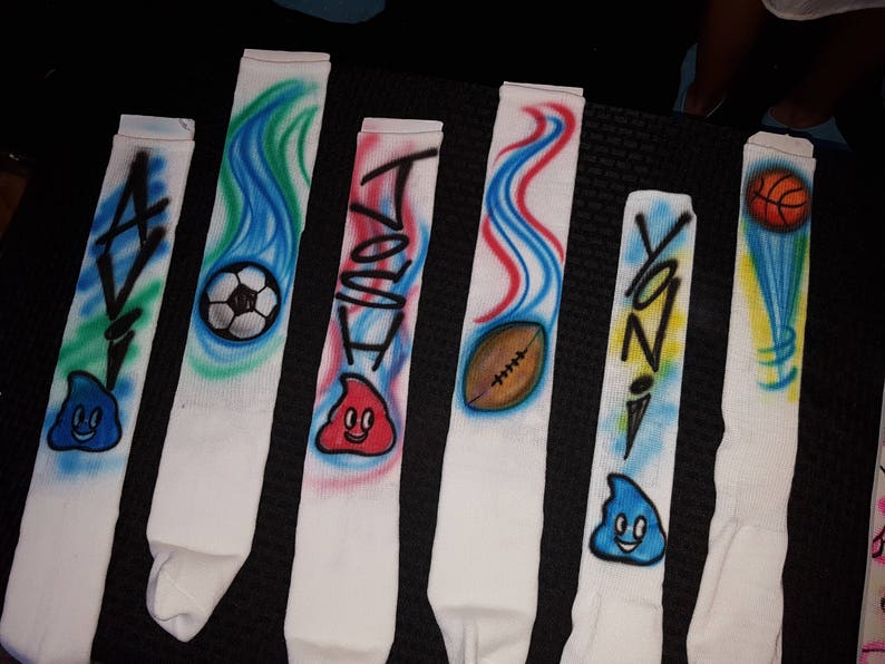 May include: White athletic socks with airbrushed designs. Each sock features a name, sports ball, or cartoon character. Designs include a soccer ball, football, basketball, and a smiling blue character. The names are in black graffiti-style lettering.