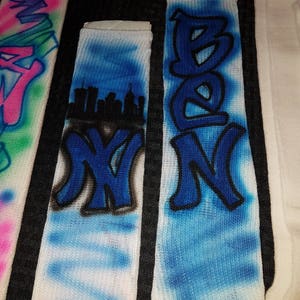 May include: White socks with graffiti-style lettering and designs. One sock features the name "BEN" in blue, another displays "NY" with a cityscape, and a third has the letter "W" in pink and blue. The socks have a spray-painted aesthetic.