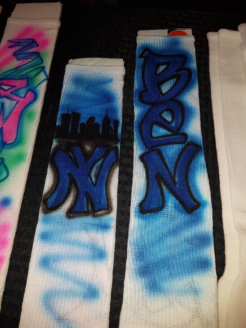May include: White socks with graffiti-style lettering and designs. One sock features the name "BEN" in blue, another displays "NY" with a cityscape, and a third has the letter "W" in pink and blue. The socks have a spray-painted aesthetic.