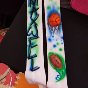 May include: Pair of white knee-high socks with airbrushed designs. One sock features the name "MAXWELL" in blue and green. The other sock has a basketball and hoop design, plus a football with a green and blue accent.