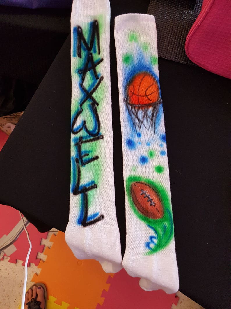 May include: Pair of white knee-high socks with airbrushed designs. One sock features the name "MAXWELL" in blue and green. The other sock has a basketball and hoop design, plus a football with a green and blue accent.