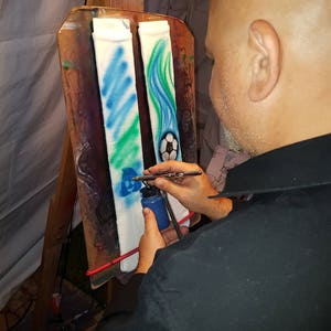 May include: A person airbrushing white socks with blue, green, and white designs. One sock features a soccer ball, while the other has a wavy pattern. The artist is using a blue paint bottle and airbrush.