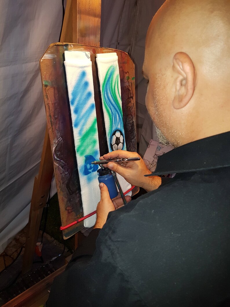 May include: A person airbrushing white socks with blue, green, and white designs. One sock features a soccer ball, while the other has a wavy pattern. The artist is using a blue paint bottle and airbrush.