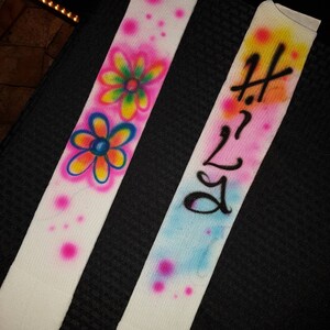 May include: Two white fabric straps with airbrushed designs. One strap features colorful flowers and pink polka dots. The other strap has the text "Hi 2 Lo" in black over a blue and yellow background with pink polka dots.