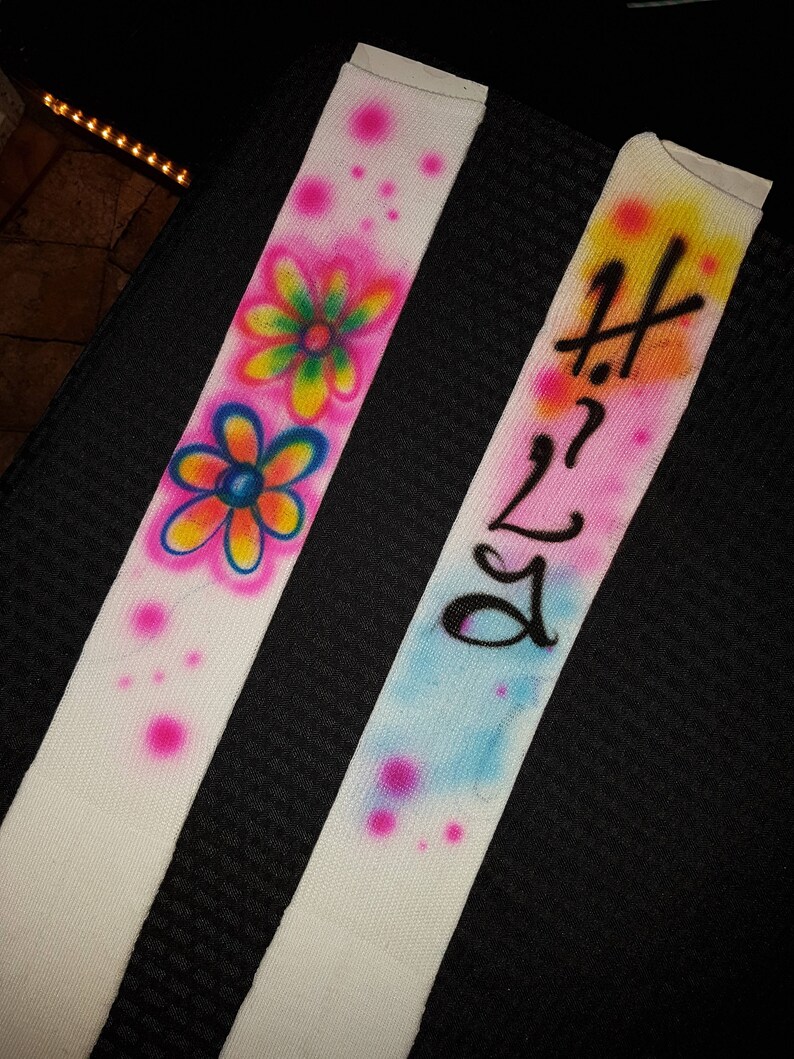 May include: Two white fabric straps with airbrushed designs. One strap features colorful flowers and pink polka dots. The other strap has the text "Hi 2 Lo" in black over a blue and yellow background with pink polka dots.
