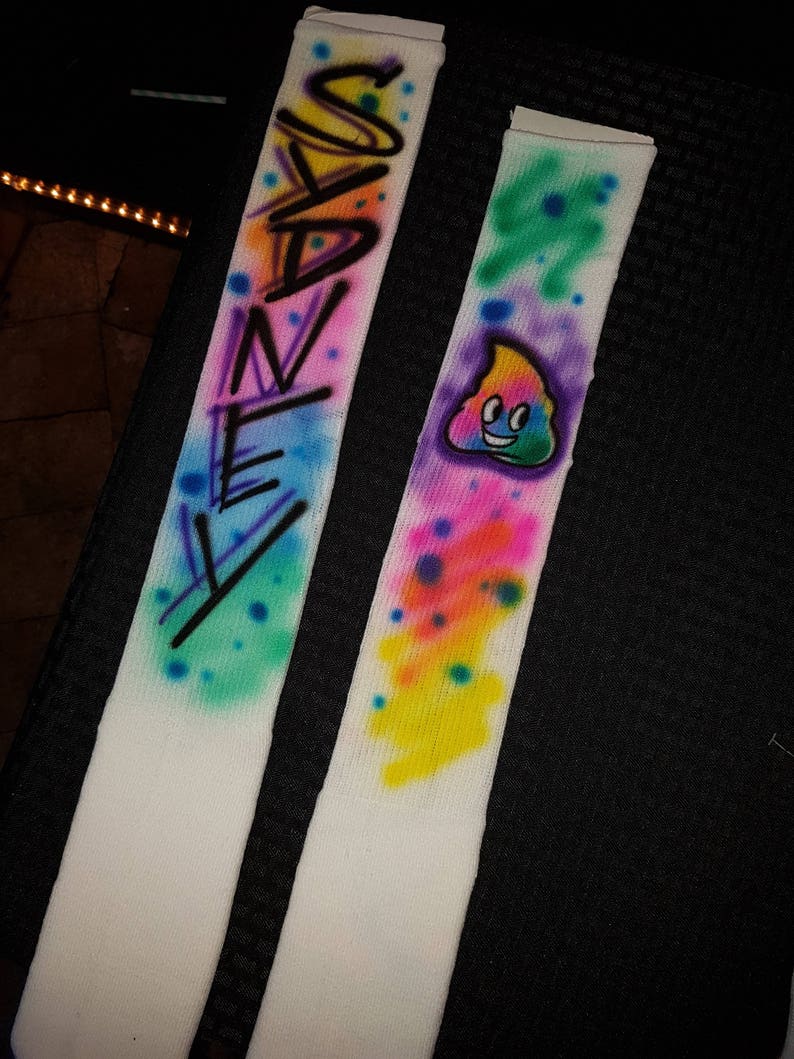 May include: Pair of white socks with airbrushed designs. One sock features the name "SANEY" in black and colorful spray paint. The other sock has a smiling poop emoji with colorful paint splatters.