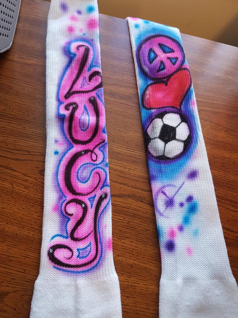 May include: Two white athletic socks with airbrushed designs. One sock features the name "LUCY" in pink and black, outlined in blue. The other sock has a peace sign, a red heart, and a soccer ball, with blue and pink accents.