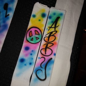 May include: White socks with airbrushed designs. One sock features a peace sign and colorful splatters. The other sock has the name "ABBY" in black script. The designs include blue, pink, yellow, and green colors.
