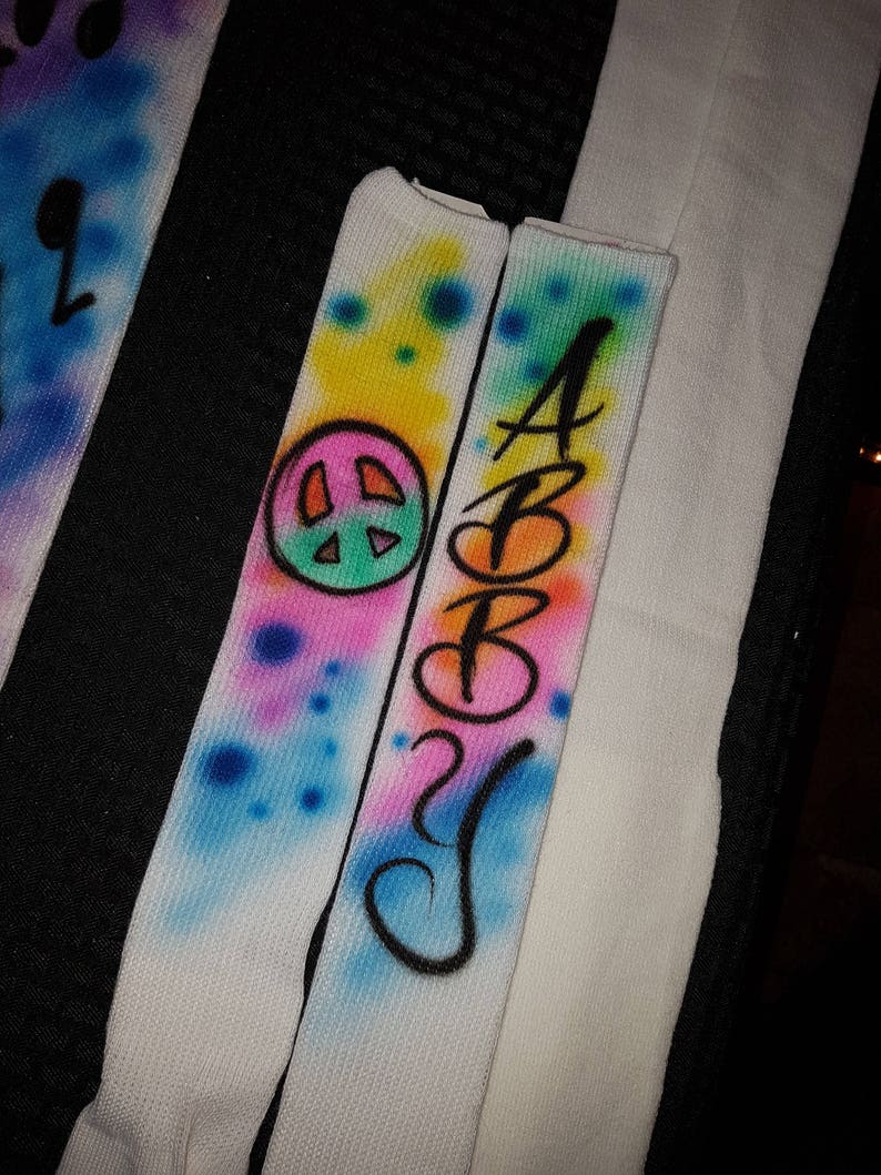 May include: White socks with airbrushed designs. One sock features a peace sign and colorful splatters. The other sock has the name "ABBY" in black script. The designs include blue, pink, yellow, and green colors.
