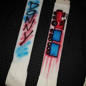 May include: Two white socks with airbrushed designs. One sock has the name "DONNY" in blue and red. The other sock features a red and black train design with blue squares. The designs are on a white ribbed fabric.