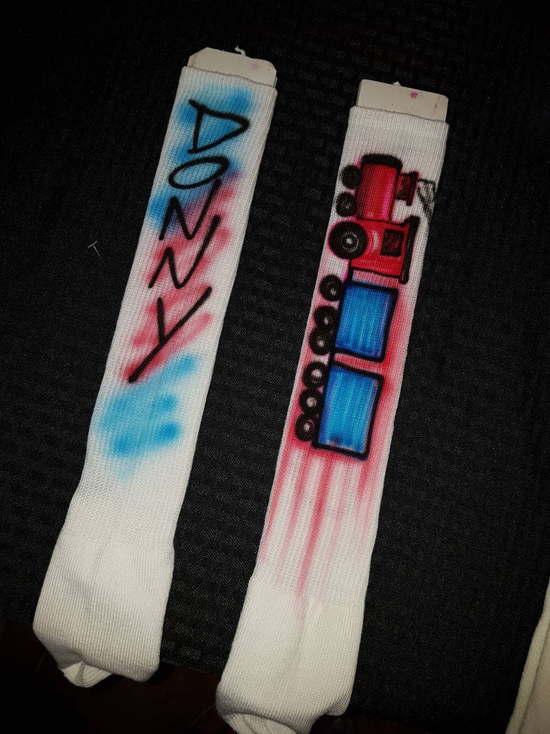 May include: Two white socks with airbrushed designs. One sock has the name "DONNY" in blue and red. The other sock features a red and black train design with blue squares. The designs are on a white ribbed fabric.