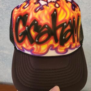 May include: Black mesh trucker hat with a black bill. The front of the hat features a white background with a yellow, orange, and purple flame design. The name "Grayson" is written in black graffiti style over the flames.