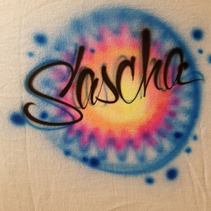 May include: A white t-shirt with a blue, pink, and yellow tie-dye design. The name "Sascha" is written in black cursive lettering over the design.