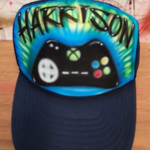 May include: A blue and green tie-dye trucker hat with the name "HARRISON" written in black across the front. A black and green video game controller is painted on the front of the hat.