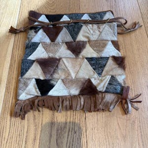 Può includere: Una pochette rettangolare realizzata con toppe triangolari di pelle bovina nei toni del marrone, nero, bianco e grigio. La borsa presenta una chiusura con coulisse in pelle marrone sulla parte superiore e frange in strisce di pelle sul fondo.