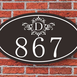 May include: Black oval house number sign with white lettering. The number is 867 and the letter D is in a decorative swirl design.