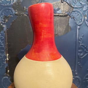 May include: A handcrafted ceramic vase with a bulbous, cream-colored base and a vibrant red neck. The vase has a smooth, glossy finish and a unique, organic shape. The vase is displayed on a wooden surface.