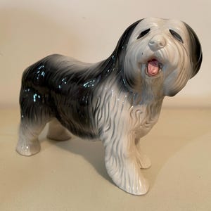 May include: A ceramic Old English Sheepdog figurine, featuring a black and white coat with a pink tongue. The dog is in a standing position, with its mouth open. The figurine is a decorative item.