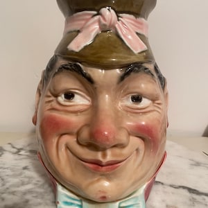 Puede incluir: Una jarra de cerámica con forma de rostro de hombre sonriente. La jarra tiene un sombrero marrón con un lazo rosa, mejillas rosadas y una pajarita azul. La cara está detallada con rasgos pintados.