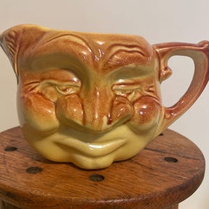 May include: A yellow and brown ceramic pitcher shaped like a face with a wide smile and closed eyes. The pitcher has a handle on the right side.