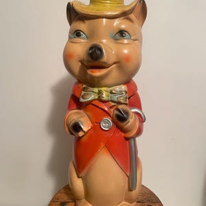 Może przedstawiać: Figurka świni w stylu vintage, w żółtym kapeluszu, czerwonym płaszczu i zielonej muszce. Świnia trzyma srebrną laskę i ma różowe policzki. Figurka stoi na drewnianej szpuli.