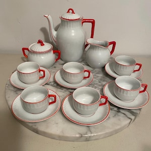 May include: A white porcelain tea set with red accents. The set includes a teapot, sugar bowl, creamer, and six teacups with saucers. The rims and handles are red, creating a striking contrast. The set is arranged on a marble surface.