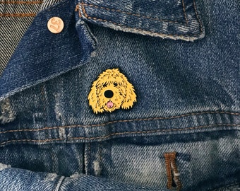 Può includere: Uno spillo dorato a forma di barboncino dorato con la lingua rosa che sporge. Lo spillo è su una giacca di jeans blu.