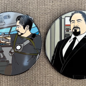 May include: Two round enamel pins featuring character portraits. One pin depicts a man in a black suit and tie. The other pin shows a man in a gray shirt operating machinery. Both pins have a silver border and are set against a neutral background.