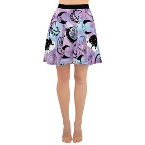 May include: A skater skirt featuring a celestial design. The skirt has a black waistband and a pattern of black moons, stars, and floral elements on a purple, blue, and white tie-dye background. The skirt is knee-length.