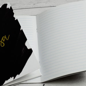May include: A black and white notebook with the word "anisa" written in gold on the front cover. The notebook is open to a blank page with lined paper.