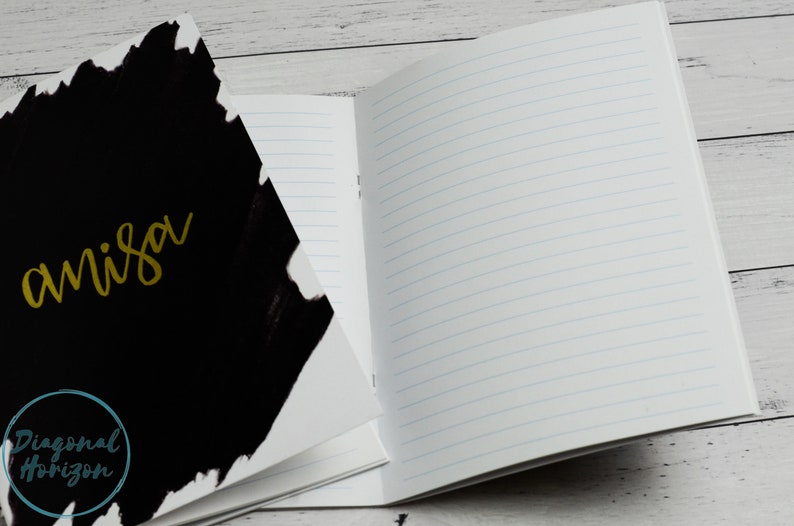 May include: A black and white notebook with the word "anisa" written in gold on the front cover. The notebook is open to a blank page with lined paper.
