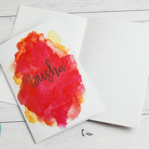 May include: A white notebook with a red, yellow and orange watercolour design on the front cover. The name "Nousha" is written in black cursive on the cover.