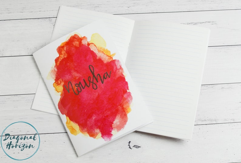May include: A white notebook with a red, yellow and orange watercolour design on the front cover. The name "Nousha" is written in black cursive on the cover.