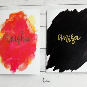May include: Two blank cards with watercolour designs. The card on the left has a red, orange, and yellow watercolour design with the name "Nousha" written in black. The card on the right has a black watercolour design with the name "amisa" written in gold.