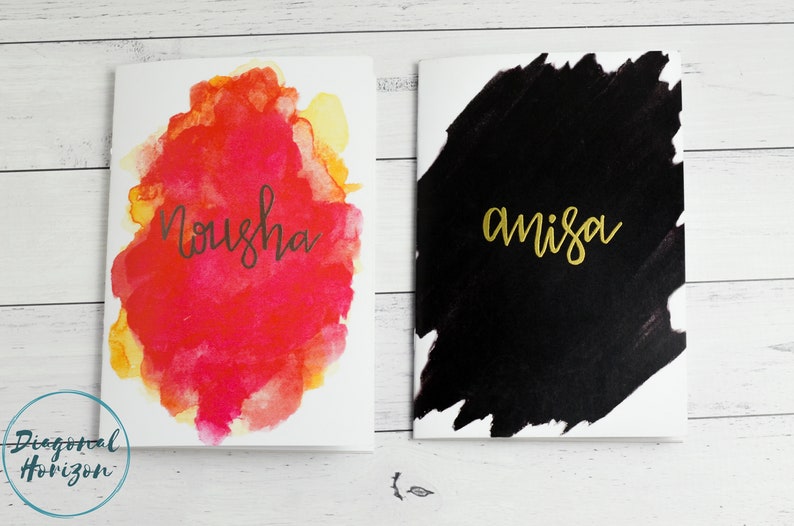 May include: Two blank cards with watercolour designs. The card on the left has a red, orange, and yellow watercolour design with the name "Nousha" written in black. The card on the right has a black watercolour design with the name "amisa" written in gold.