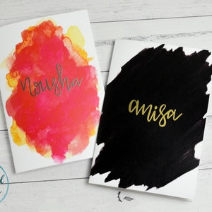 May include: Two blank greeting cards with watercolour designs. The card on the left has a pink and yellow watercolour design with the name "Nousha" written in silver lettering. The card on the right has a black watercolour design with the name "amisa" written in gold lettering.