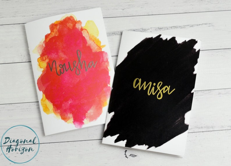 May include: Two blank greeting cards with watercolour designs. The card on the left has a pink and yellow watercolour design with the name "Nousha" written in silver lettering. The card on the right has a black watercolour design with the name "amisa" written in gold lettering.