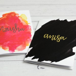 May include: Two small notebooks with watercolour painted covers. The first notebook has a pink and yellow watercolour design with the word "Nousha" written in black. The second notebook has a black watercolour design with the word "amisa" written in gold.
